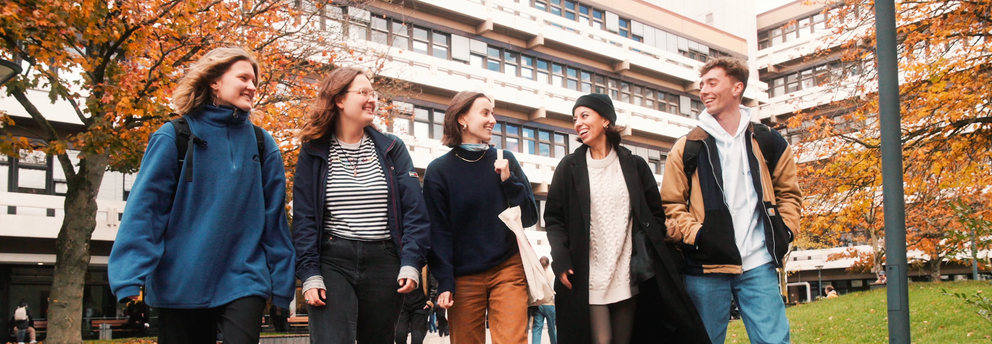 Header image IJ students autumn EF50 5 students, male and female, are walking on the campus in front of EF 50 building, they are happily chatting, the brownish leaves on the trees and the student's clothing suggest that it's autumn